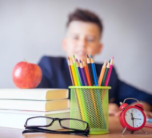 Zdjęcie przedstawia biurko ucznia w szkole. Na biurku znajdują się kredki, książki, jabłko, okulary i zegar. W tle widać sylwetkę chłopca.